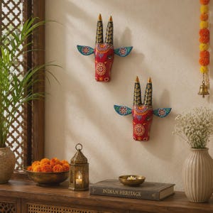 May include: Two hand-painted wall sculptures of bull heads, each featuring a vibrant red face with intricate white and yellow floral patterns, black horns with gold tips, and blue ears. The bull heads are mounted on a plain off-white wall surface.