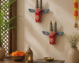 May include: Two hand-painted wall sculptures of bull heads, each featuring a vibrant red face with intricate white and yellow floral patterns, black horns with gold tips, and blue ears. The bull heads are mounted on a plain off-white wall surface.
