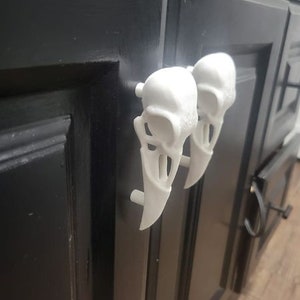 Peut inclure: Deux poignées de porte de placard en forme de crâne blanc. Les poignées sont fixées à une porte de placard noire.
