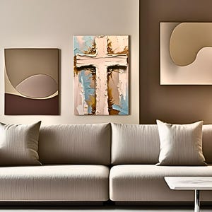 May include: An interior design scene featuring a beige sofa with two throw pillows and a white side table. Above the sofa, three pieces of wall art are displayed. The central artwork is a textured painting of a cross with blue, brown, and white brushstrokes.