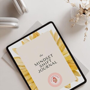 May include: A digital tablet displaying a journal cover with a watercolor sunflower design. The cover text reads "the MINDSET SHIFT JOURNAL" and features a pink circle with the text "YOUR THERAPY PARTNER".