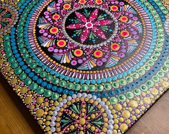 May include: A colorful mandala painting on canvas, created using the dot painting technique. The design features a central flower with multiple layers of concentric circles in shades of pink, green, blue, gold, and silver.
