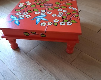 May include: A small orange wooden table with hand-painted floral designs and two blue birds. The table has four legs and is sitting on a wooden floor.