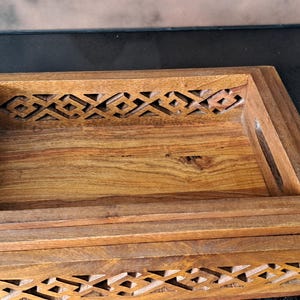 以下が含まれることがあります： 複雑な彫刻が施された木製のトレイ3個セット。トレイは互いに重ねて収納でき、薄い茶色の木でできています。