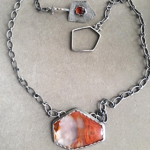 Peut inclure: Un collier en argent avec trois pendentifs. Le plus grand pendentif présente une grande pièce d'agate de forme irrégulière avec des bandes rouges, orange et blanches. Les deux autres pendentifs sont en argent et de forme géométrique. L'un des plus petits pendentifs a une petite pierre rouge.