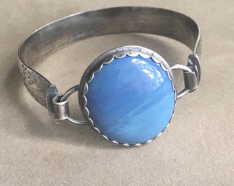 May include: A silver-toned bracelet with a large, round, light blue centerpiece. The centerpiece is framed by a scalloped edge. The bracelet band has a hammered texture on one side and connects with a simple clasp.