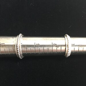 Pu&ograve; includere: Due anelli d'argento con un design a perline sono esposti su un misuratore di anelli. Gli anelli sono posizionati sui segni di 15 e 18 cm. Lo sfondo &egrave; di colore scuro e uniforme.