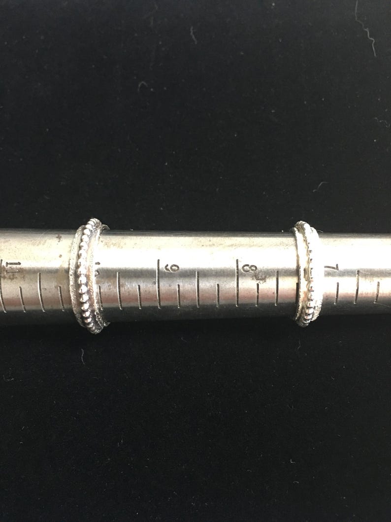 Pu&ograve; includere: Due anelli d'argento con un design a perline sono esposti su un misuratore di anelli. Gli anelli sono posizionati sui segni di 15 e 18 cm. Lo sfondo &egrave; di colore scuro e uniforme.