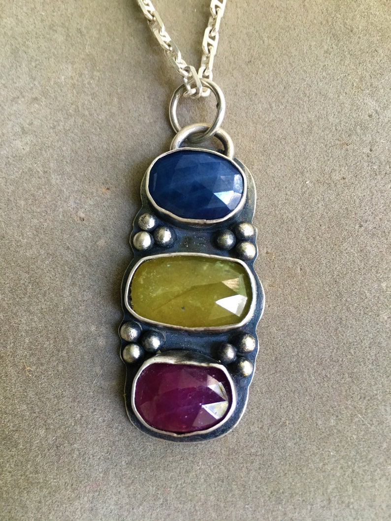May include: A silver pendant necklace featuring three colorful gemstones: a deep blue sapphire, a yellow-green gemstone, and a vibrant ruby. The pendant is suspended from a silver chain, showcasing a unique and eye-catching design.