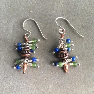 Può includere: Orecchini fatti a mano con ganci in argento, filo di rame e perline decorative. Gli orecchini hanno un design unico con perline blu, verdi e color rame raggruppate. Gli orecchini sono progettati per dondolare.