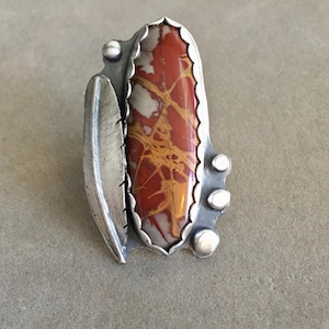 May include: A silver ring featuring a large, oval-shaped stone with a red, brown, and cream-colored pattern. The stone is set in a scalloped bezel. A silver leaf shape and three silver beads adorn the ring's design.