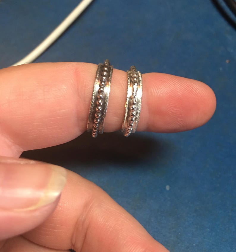 Pu&ograve; includere: Due anelli d'argento con un design a perline sono mostrati su un dito. Gli anelli hanno una fascia strutturata con una fila di piccole perline in rilievo lungo il centro. Sono gioielli.