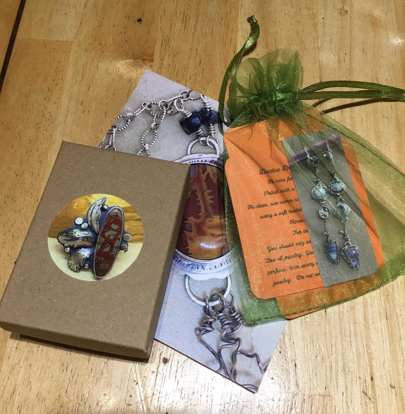 May include: An assortment of handmade jewelry, including a ring in a brown box, a necklace, and a pair of earrings in a green mesh bag. The ring features a large, oval, reddish-brown stone. The earrings have blue and clear stones.