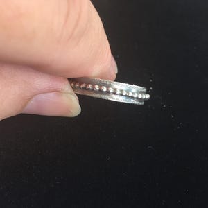 Pu&ograve; includere: Un anello d'argento con una fascia strutturata e una fila di piccole perline rialzate su entrambi i lati. L'anello &egrave; tenuto su uno sfondo scuro, mettendo in risalto il suo design e i dettagli.