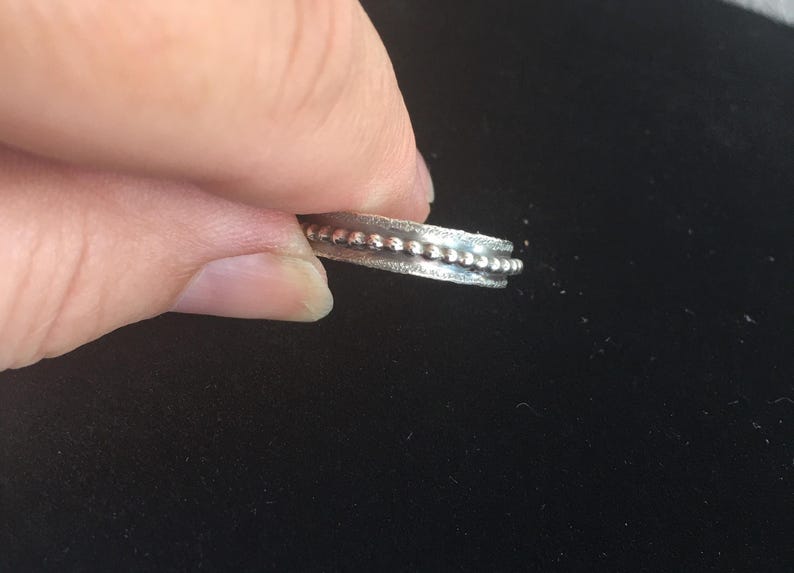 Pu&ograve; includere: Un anello d'argento con una fascia strutturata e una fila di piccole perline rialzate su entrambi i lati. L'anello &egrave; tenuto su uno sfondo scuro, mettendo in risalto il suo design e i dettagli.