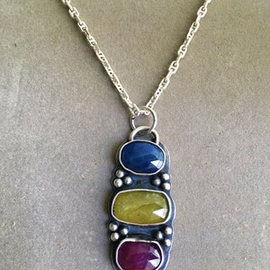 May include: A silver necklace with a pendant featuring three oval-cut gemstones in blue, yellow-green, and red-purple. The pendant is rectangular with a textured border and small silver beads. The chain is a delicate silver link.