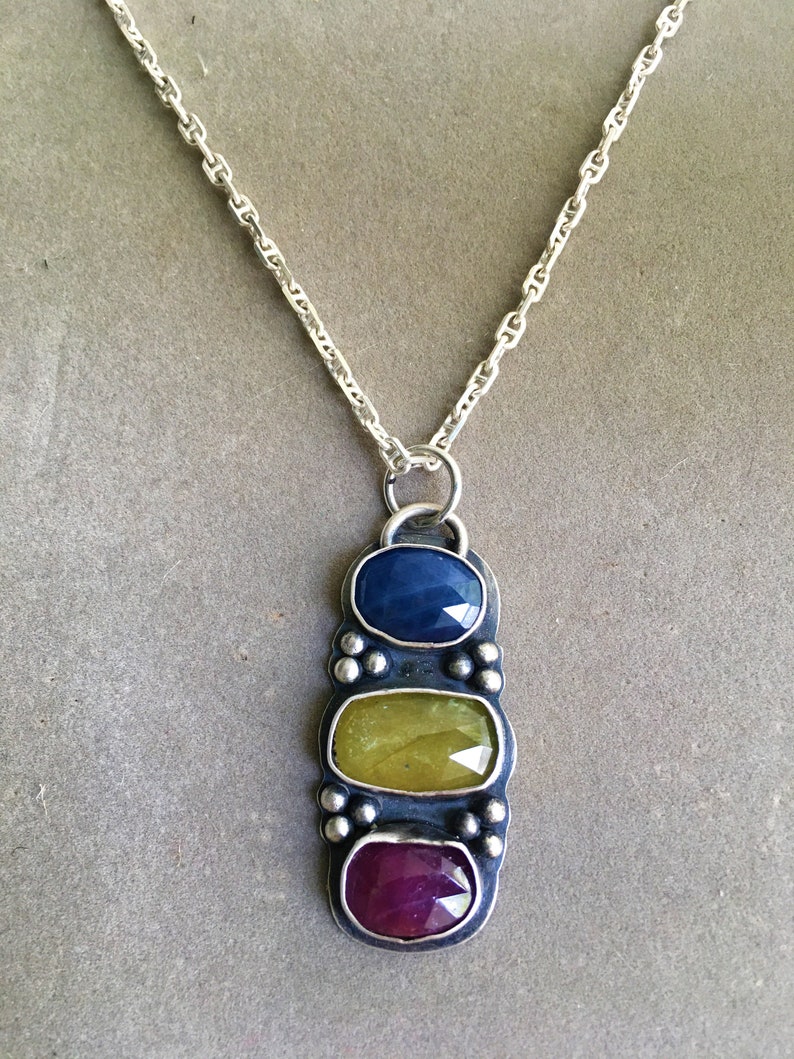 May include: A silver necklace with a pendant featuring three oval-cut gemstones in blue, yellow-green, and red-purple. The pendant is rectangular with a textured border and small silver beads. The chain is a delicate silver link.