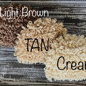 May include: Three fluffy yarn balls in different shades of brown: light brown, tan, and cream. The yarn balls are arranged in a row on a wooden surface.