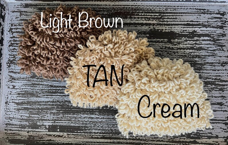 May include: Three fluffy yarn balls in different shades of brown: light brown, tan, and cream. The yarn balls are arranged in a row on a wooden surface.