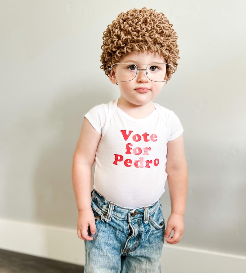 May include: A young child wearing a white bodysuit with the text "Vote for Pedro" in red and pink letters. The child is also wearing a brown curly wig and glasses. The child is wearing blue jeans.