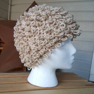 May include: A beige crocheted hat with a curly texture. The hat is on a white mannequin head.