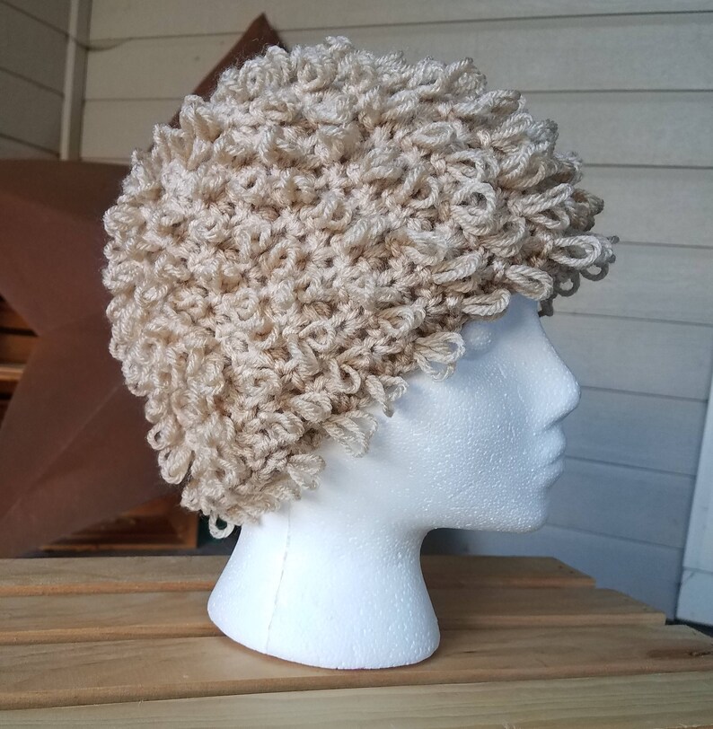 May include: A beige crocheted hat with a curly texture. The hat is on a white mannequin head.