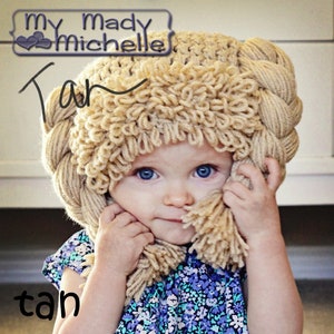 May include: A young child wearing a tan crocheted hat with a braided design. The hat has a fluffy texture and is shaped like a crown. The child is wearing a blue and purple floral print shirt.