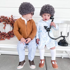 May include: Two children dressed as elderly people for Halloween. One child is wearing a brown sweater, blue pants, and a gray wig. The other child is wearing a blue dress, white socks, brown shoes, and a gray wig. They are sitting on a white bench with a vintage rotary phone.
