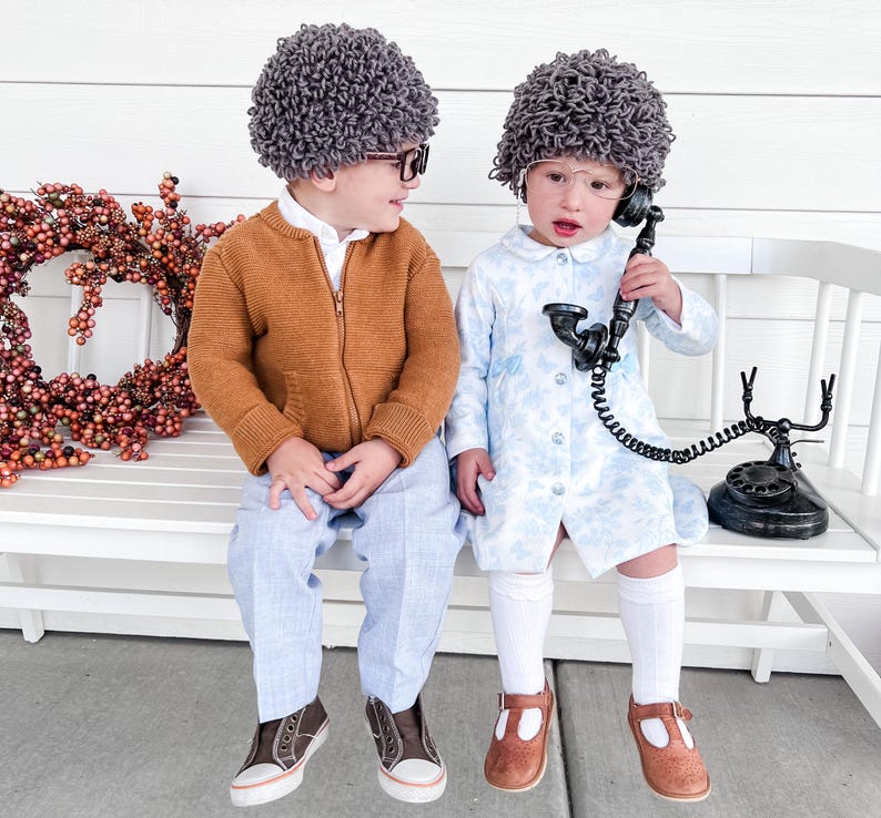 May include: Two children dressed as elderly people for Halloween. One child is wearing a brown sweater, blue pants, and a gray wig. The other child is wearing a blue dress, white socks, brown shoes, and a gray wig. They are sitting on a white bench with a vintage rotary phone.