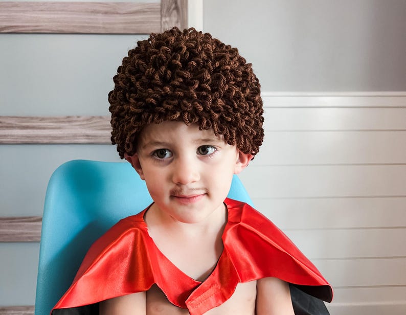 May include: A young child wearing a brown curly wig and a red cape, sitting on a blue chair.