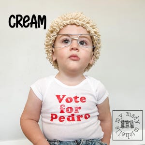 May include: A child wearing a white bodysuit with the text "Vote for Pedro" in red letters. The child is wearing a cream-colored wig and glasses.