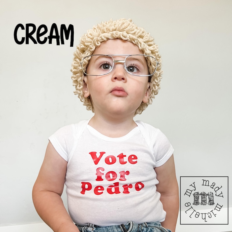 May include: A child wearing a white bodysuit with the text "Vote for Pedro" in red letters. The child is wearing a cream-colored wig and glasses.