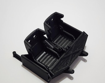 May include: Black plastic model car seats with a detailed textured design. The seats are connected and have a small metal rod attached to the side.