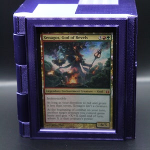 May include: A purple card holder with a framed Magic: The Gathering card. The card features the text "Xenagos, God of Revels" and a colorful illustration of a mythical figure. The card is 6/5.