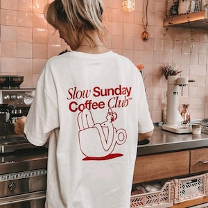 May include: White t-shirt with a red graphic of a person relaxing in a coffee cup. The text "Slow Sunday Coffee Club" is printed in red on the back of the shirt.