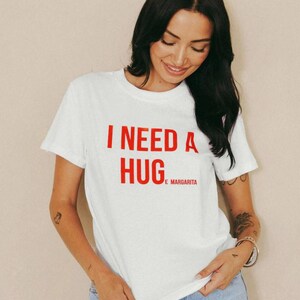 May include: White t-shirt with the red text "I NEED A HUG" and "E MARGARITA" underneath. The shirt has short sleeves and a crew neck. The model is wearing blue jeans.