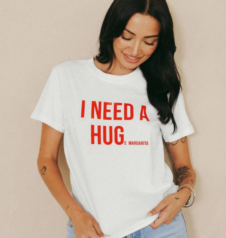 May include: White t-shirt with the red text "I NEED A HUG" and "E MARGARITA" underneath. The shirt has short sleeves and a crew neck. The model is wearing blue jeans.