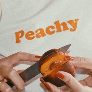 May include: A person is cutting a peach in half with a silver knife. The peach is ripe and juicy, with a bright orange flesh. The person is wearing a white shirt with the word "Peachy" printed in orange letters.