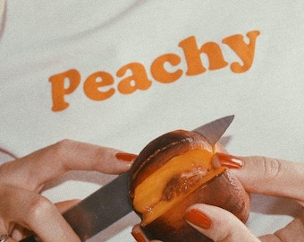 Op de afbeelding: Een persoon snijdt een perzik doormidden met een zilveren mes. De perzik is rijp en sappig, met een helder oranje vruchtvlees. De persoon draagt een wit shirt met het woord "Peachy" in oranje letters.
