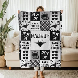 Puede incluir: Una manta de patchwork en blanco y negro con gafas de aviador, aviones de combate, cascos de piloto y el texto "TALK TO ME GOOSE" y "MAVERICK". La manta se muestra frente a un sofá beige.