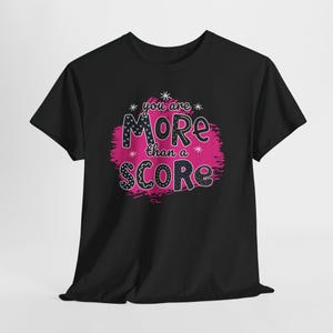 May include: Black t-shirt with the phrase "you are MORE than a SCORE" in white, black, and pink lettering. The word "MORE" has a pattern of black and white polka dots and stars. The text is on a pink background.