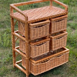 May include: A light brown wicker storage unit with five woven baskets. The unit has a rectangular top and a sturdy frame. The baskets are of varying sizes, with the largest at the bottom. The unit is set on a grassy surface.