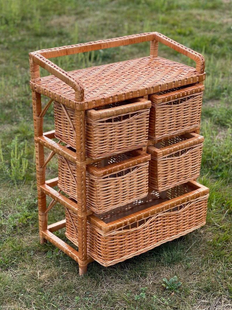 May include: A light brown wicker storage unit with five woven baskets. The unit has a rectangular top and a handle. The baskets are of varying sizes and are stacked on top of each other. The unit is made of woven wicker.
