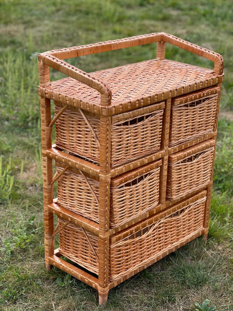 May include: A light brown wicker storage unit with six drawers and a top shelf. The unit is made of woven wicker and has a rectangular shape. The drawers have curved handles and the unit is on a grassy surface.