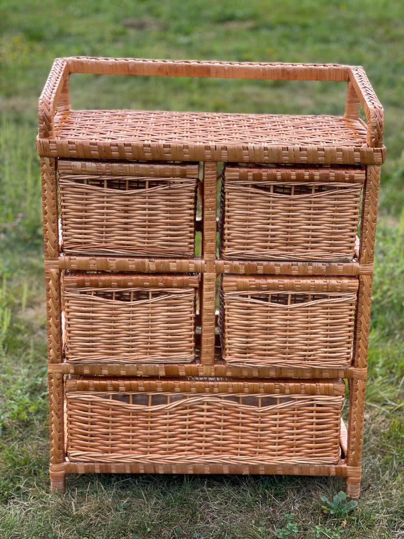 May include: A light brown wicker storage unit with five woven baskets. The unit has a rectangular shape with a handle on top. The baskets are of varying sizes, suitable for organizing items. The unit is made of natural materials.