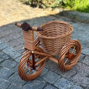 Op de afbeelding: Een decoratieve mand in de vorm van een driewieler, gemaakt van geweven wilgenhout. De driewieler heeft drie wielen, een mand voor opslag en een stuur. De natuurlijke bruine kleur van het wilgenhout geeft het een rustieke uitstraling. De driewieler staat op een stenen oppervlak.
