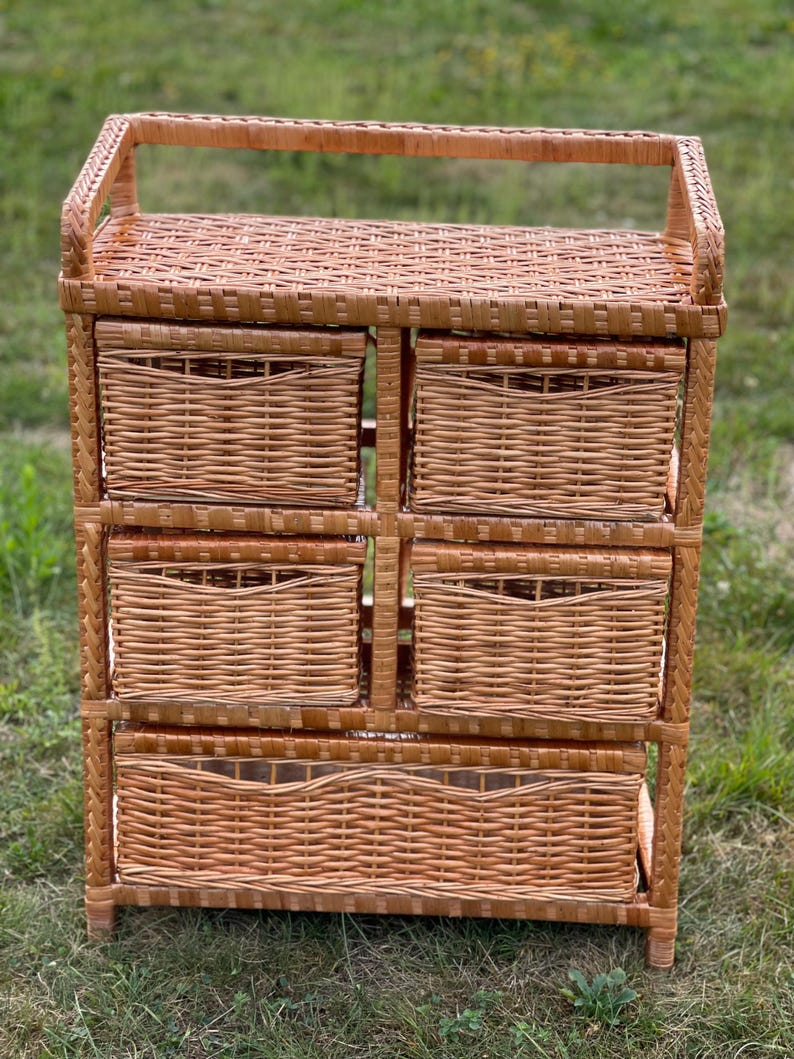 May include: A light brown wicker storage unit with six woven drawers. The unit has a rectangular shape with a handle on top. The drawers are evenly spaced and the unit is sitting on a grassy surface.