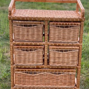 May include: A light brown wicker storage unit with five woven baskets. The unit has a rectangular shape with a handle on top. The baskets are of varying sizes, suitable for organizing items. The unit is made of natural materials.
