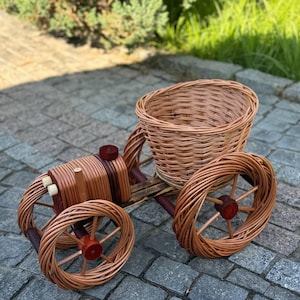 Op de afbeelding: Een handgemaakte miniatuur rieten wagen met een mand en vier wielen. De wagen is gemaakt van lichtbruin geweven riet en heeft een rustieke, natuurlijke uitstraling. De wielen en andere details zijn van hout.