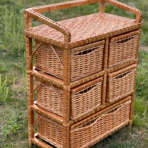 May include: A light brown wicker storage unit with six drawers. The unit has a rectangular shape with a woven top and a handle. The drawers are also woven and have a curved handle opening. The unit is made of natural wicker.